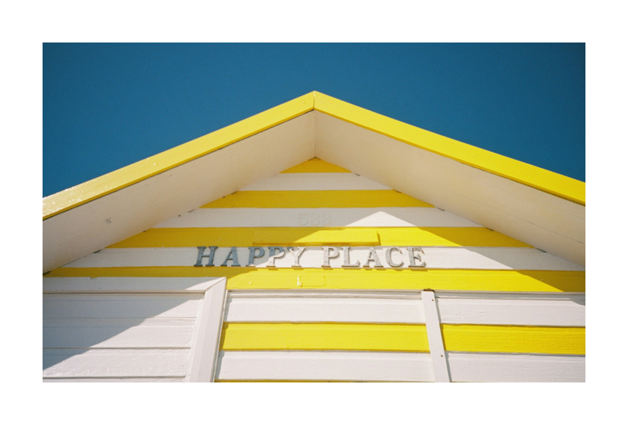 Pictured, a birds eye view of a bright yellow and white striped beach house. In the background is a beautiful blue, clear sky.
