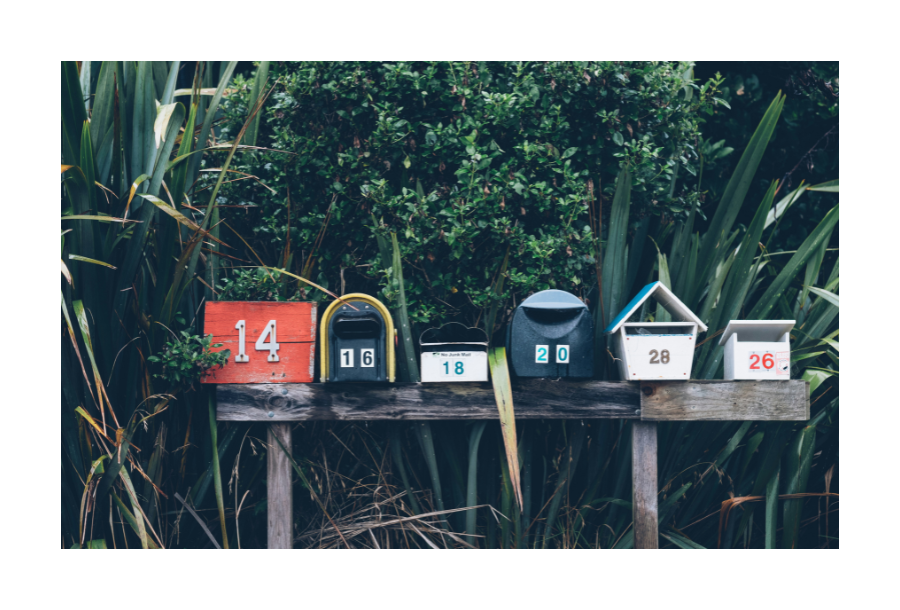 Pictured, a darker afternoon mood with dark green bushes in the background, in the foreground are six different sized, shaped and colour mail boxes all sharing their own individual character.