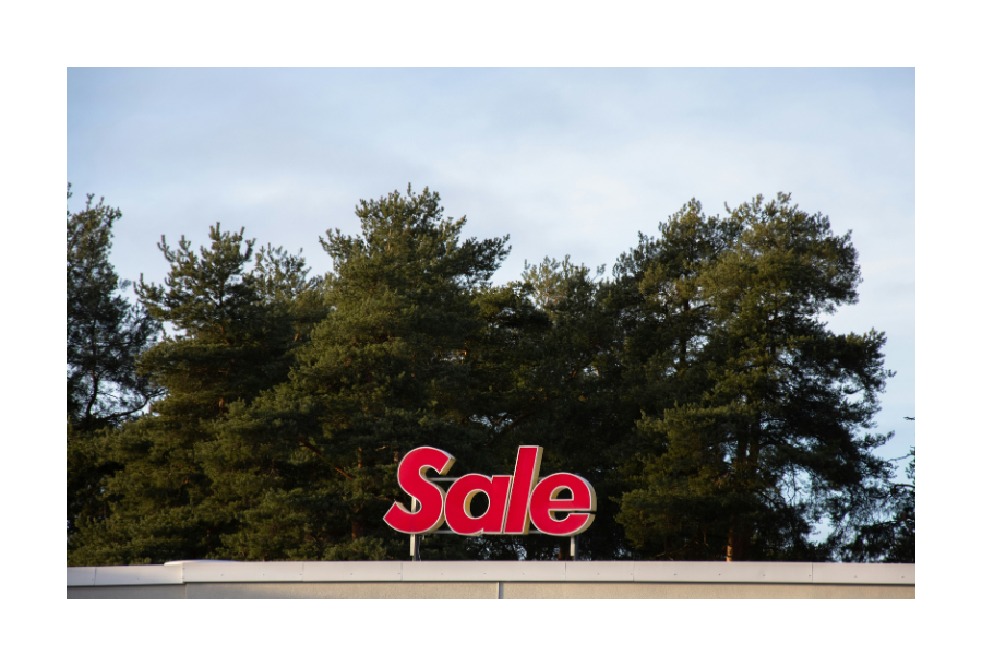 Pictured, a red "sale" sign that is secured to the top of a build. In the background a green pine trees with a cloud background.