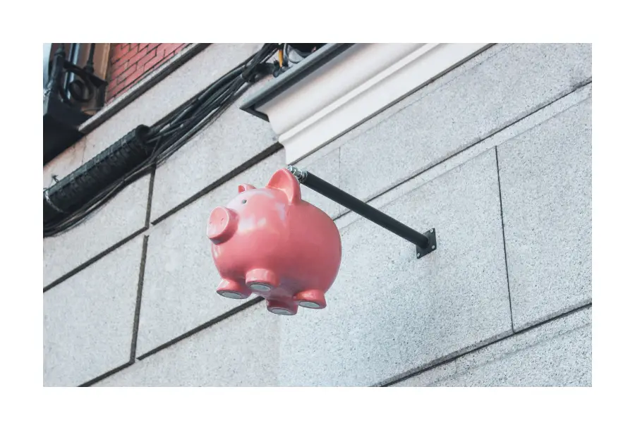 Pictured, a light brick building, zoomed in on a hanging pink piggy bank from outside of the wall.